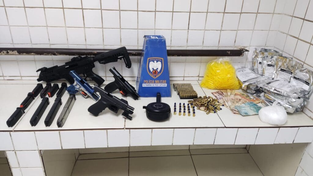 Foto de materiais apreendidos pela Polícia Militar, sobre uma bancada na Delegacia Regional situada na Serra, três pistolas de diferentes marcas e modelos, todas calibre 9mm, acessórios que aumentam o potencial ofensivo das armas, treze carregadores de pistola, 242 munições de calibre 9mm, sete munições de calibre .40, além de 500 gramas de cocaína e substâncias comumente usadas para refino de cocaína além de R$ 1.879,00 reais em espécie.