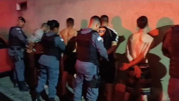 Polícia Militar prendendo 5 sujeitos encostados em um muro, todos os sujeitos estão com as mãos para trás enquanto são algemados pela PM