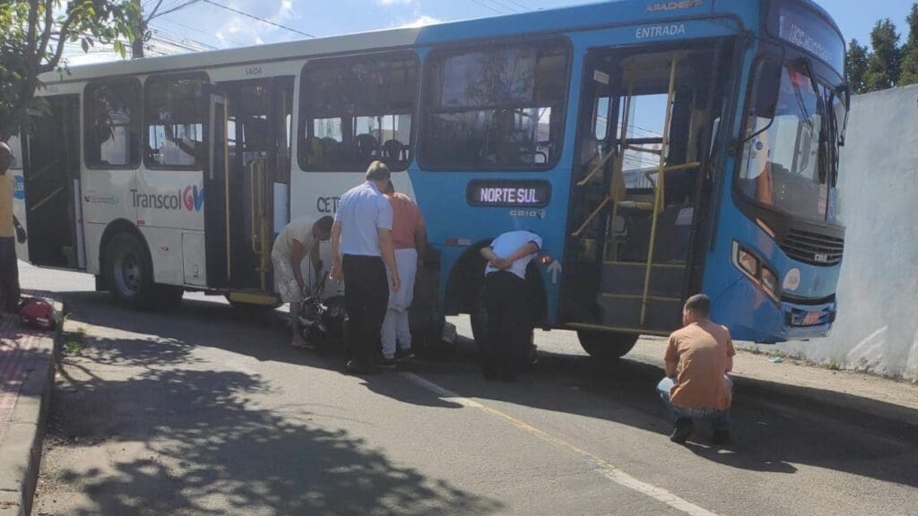 Foto com um ônibus atravessado na pista e uma motocicleta presa de baixo do veículo, envolta alguns moradores e o motorista tentando tirar a motocicleta de baixo do veículo.