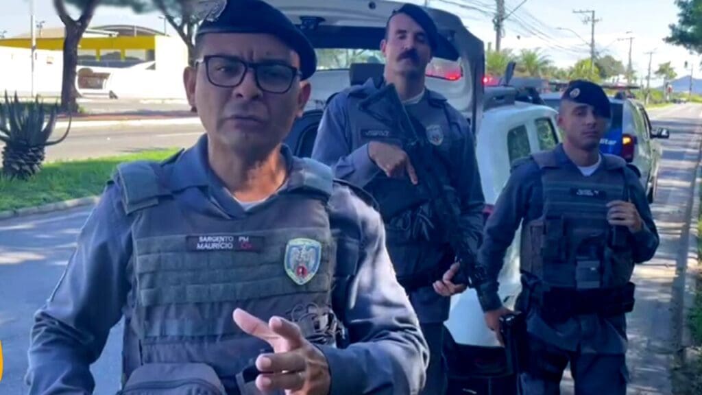 Foto com imagem de 3 policiais militares em frente a uma viatura da PM, em uma avenida movimentada de Jacaraípe