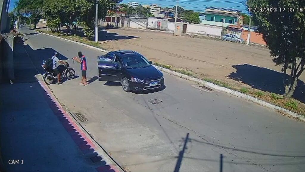 Foto da camera de segurança do momento em que um homem encapuzado armado com uma submetralhadora rende um motociclista, em uma rua deserta em plena luz do dia, e um veiculo parado com a porta do passageiro aberta.