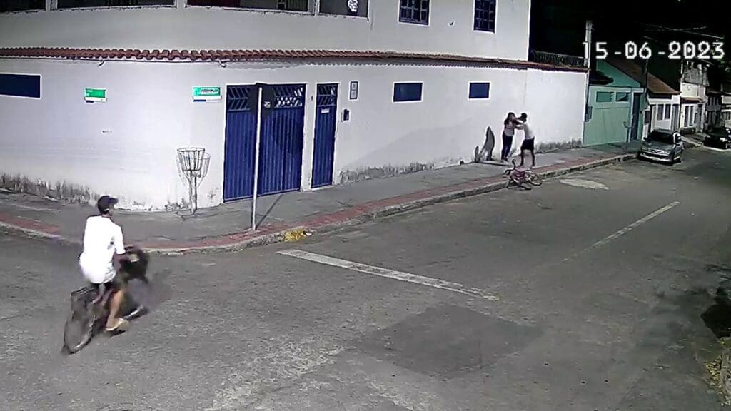 Imagem de camera de segurança mostrando uma rua com dois jovens em bicicletas assaltando uma adoescente