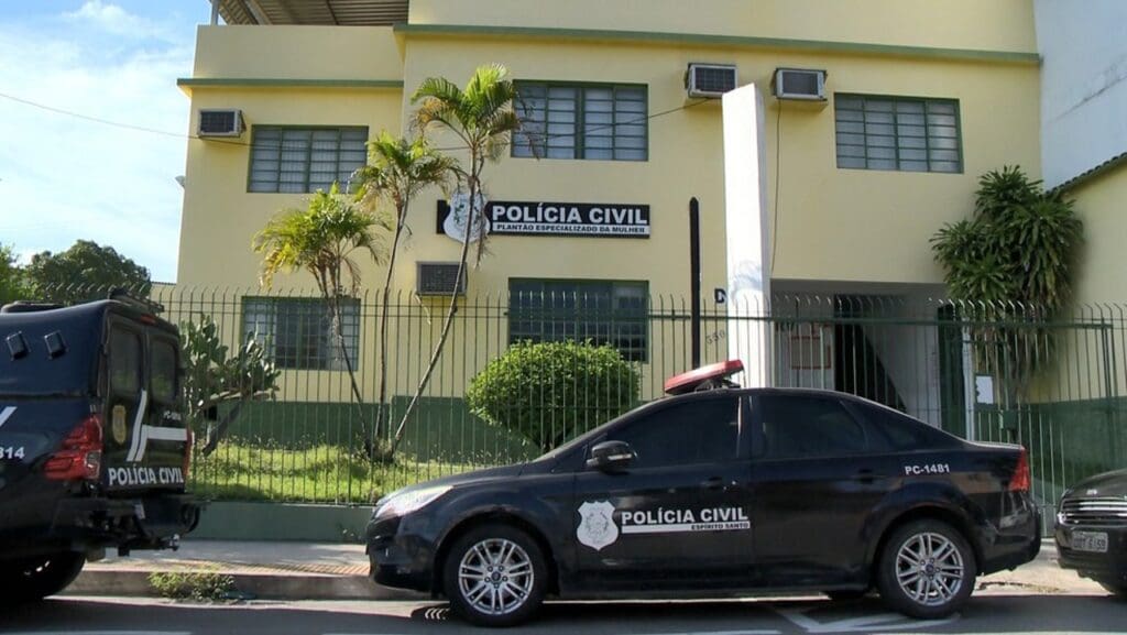 Foto da entrada da Delegacia especializada em atendimento a Mulheres, Plantão Especializado da Mulher (PEM). Um prédio de dois andares cercado por uma grade, na frente do prédio duas viatura da Policia Civil e um carro de passeio.