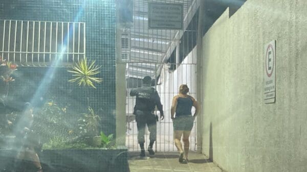 Policial Militar levando uma mulher acusada de falsificar documentos, ambos de costas em frente em frente a um portão da 3ª Delegacia Regional da Serra.