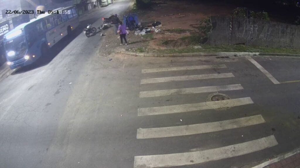 Imagens de uma câmera de segurança em um cruzamento logo após um assaltante sofrer um acidente de moto e ficar caído na calçada, na imagem há um ônibus passando pelo local e algumas pessoas em volta da vitíma.