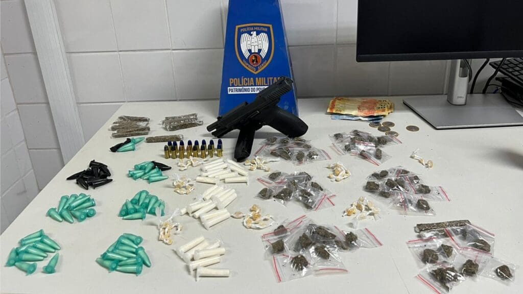 Foto de apreensão da PM, sobre uma mesa branca uma pistola 9mm, com numeração raspada, um carregador e 16 munições do mesmo calibre, além de 95 pinos de cocaína, 73 pedras de crack, 71 buchas de maconha e a quantia de R$ 181,00 em dinheiro