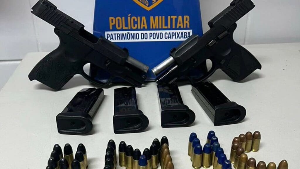 Sobre uma mesa branca uma propaganda da Polícia Militar, feita de papelão, duas pistolas 9 mm, 4 carregadores e 56 munições sendo que 45 delas são calibre 9mm e 11 são munições calibre 380.