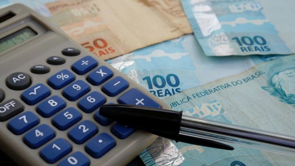 Como saber se você caiu na malha fina do Imposto de Renda 2023 Foto de uma calculadora, em cima de uma quantidade bacana de dinheiro e uma caneta bic de cor preta.