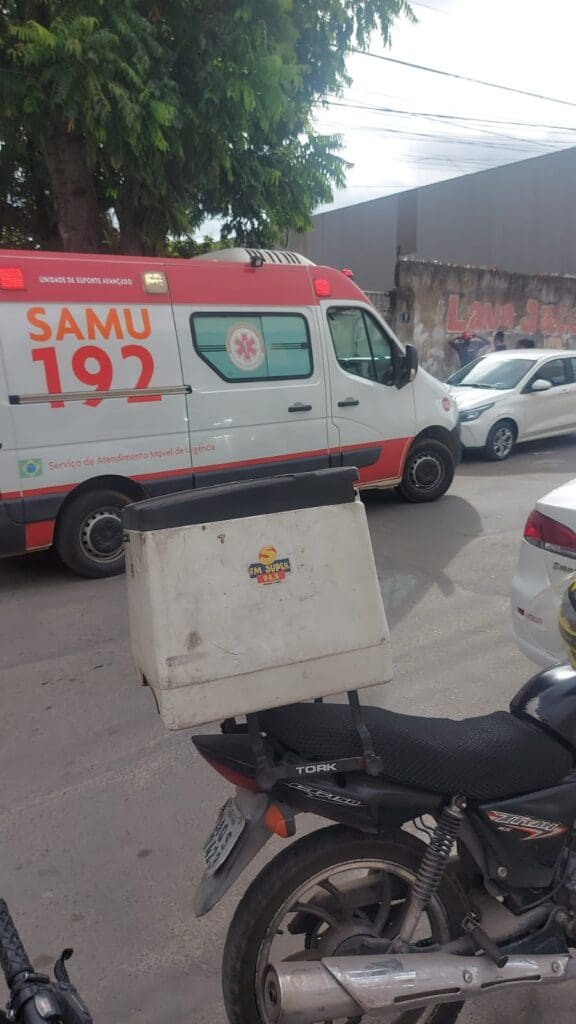 Foto de uma ambulância do SAMU estacionada em uma rua, no local há mais dois carros e uma motocicleta estacionada, além de 3 pessoas observando as moviemntações.