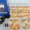 Foto de materiais apreendidos durante patrulhamento da PM, sobre uma mesa branca, um revólver calibre .38, 3 pinos de cocaína, além de dezenas de notas contabilizando 1.130 reais