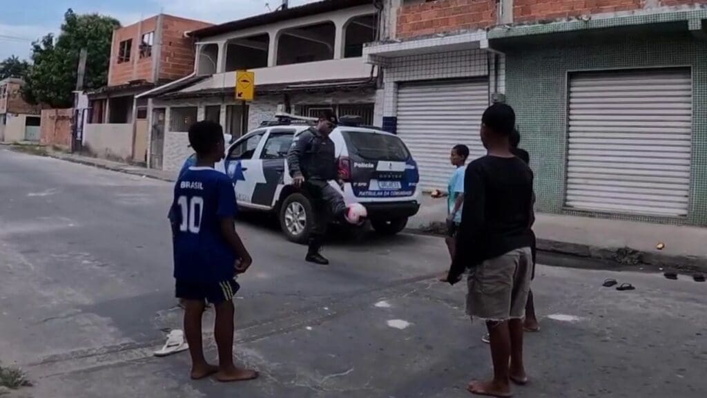 Cabo da Polícia Militar, ao lado de uma viatura da PM, com 5 crianças jogando altinha em uma residencial da Serra