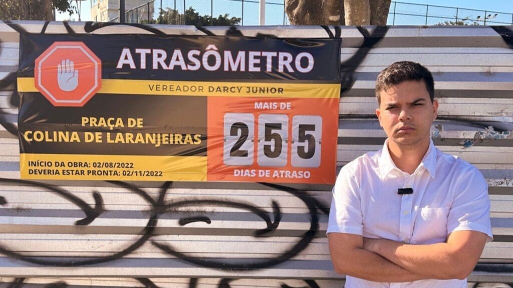 Vereador Darcy Junior de braços cruzados, em frente a uma chapa de ferro que está cercando uma praça em obras com o atraso de 255 dias, em sinal de indignação ele colou uma placa informando o atraso.