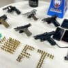 Sobre uma mesa redonda, um revólver calibre .38, 5 pistolas, 7 celulares, dezenas de munições e drogas.