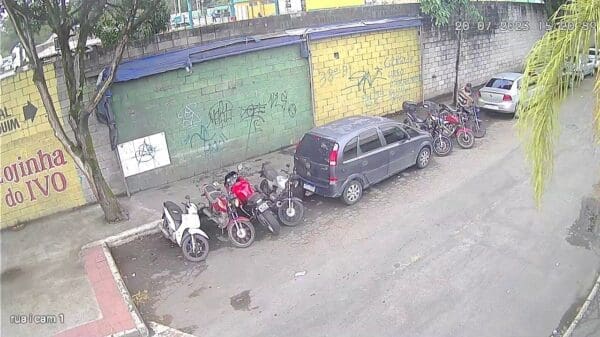 Foto de uma rua tranquila, com 10 motocicletas estacionadas proximo a guia, enquanto um ladrão força a ignição para roubar o veiculo.