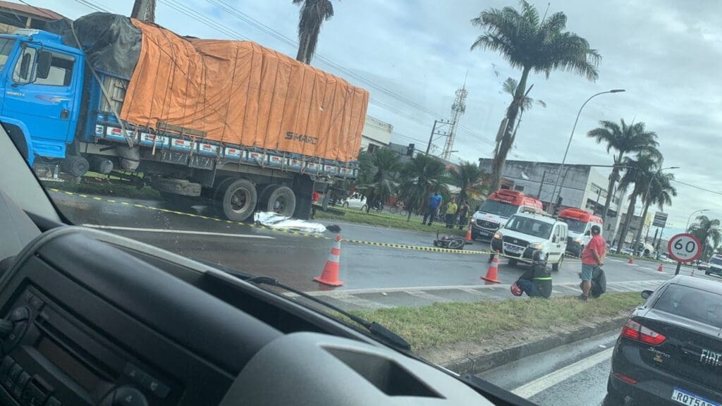 Foto da BR-101 interditada, com um caminhão estacionado na via que está molhada devido às chuvas no local, com um corpo ao lado do veículo coberto por uma manta da Polícia Civil, parados proximo ao caminhão estão a motocicleta destruída e duas ambulâncias.