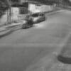Foto preto e branco de uma rua residencial, com um automóvel estacionado, e um ladrão pilotando uma moto, parado esperando seu comparsa realizar o furto de outra motociclieta.