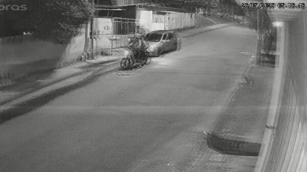 Foto preto e branco de uma rua residencial, com um automóvel estacionado, e um ladrão pilotando uma moto, parado esperando seu comparsa realizar o furto de outra motociclieta.