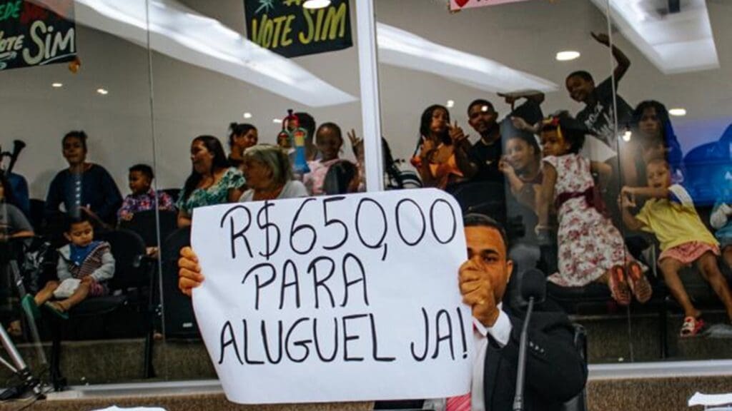 Valor do Aluguel Social na Serra sobe para 650 reais após pressão de vereador e famílias Vereador Anderson Muniz segurando uma faixa no plenário da Câmara da Serra.