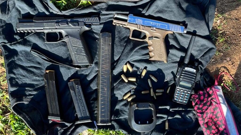 Sobre o chão forrado com um tecido, duas pistolas, 3 carregadores, um radio comunicador, uma base de recarregar os radios além de munições calibre 9mm.