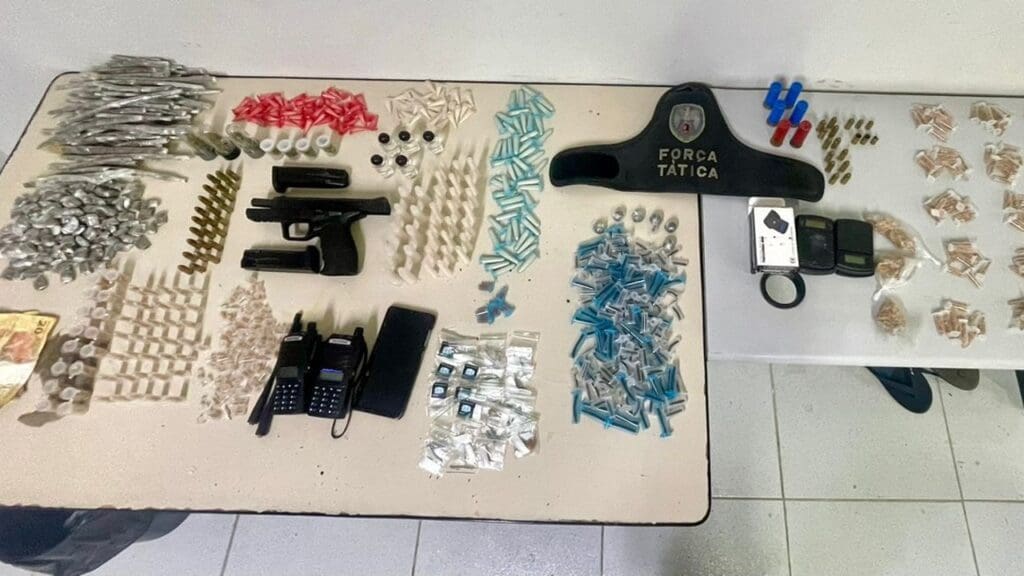 Polícia Militar apresentando, apreensões realizadas em ações nos bairros de Vitória. Sobre uma mesa centenas de apreensões de drogas, e armas.