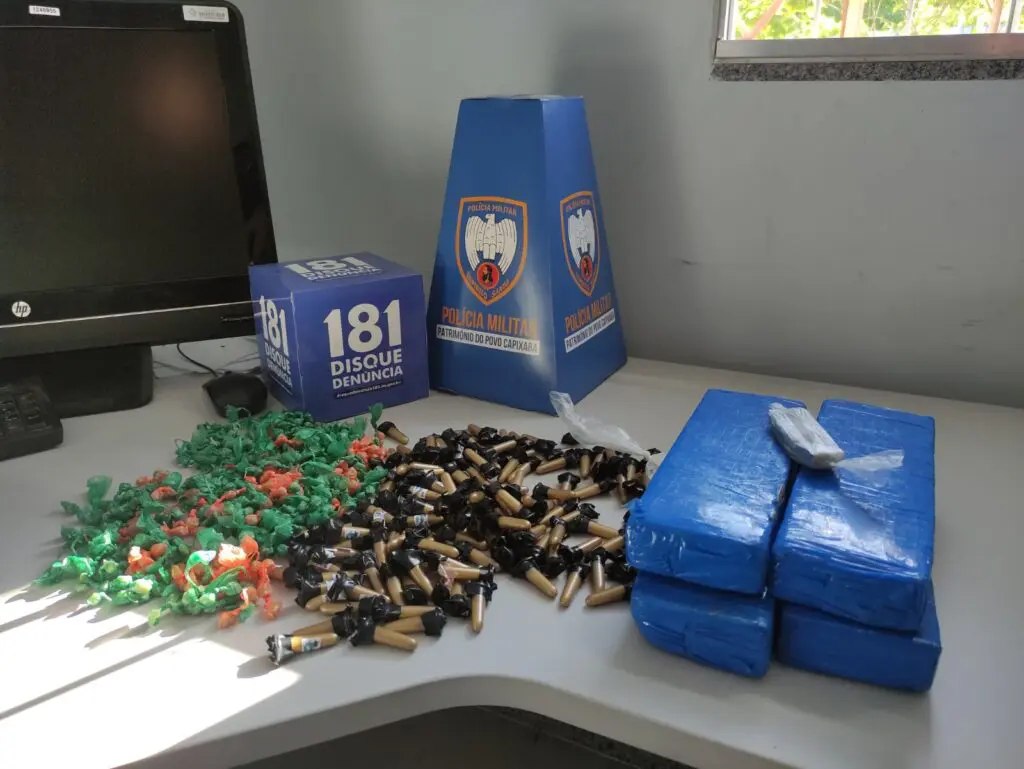 Apreensão realizada pela equipe da PM, sobre uma mesa 572 pedras de crack, 140 recipientes com cocaína e quatro pacotes consideráveis de maconha, juntamente com itens para embalagem.