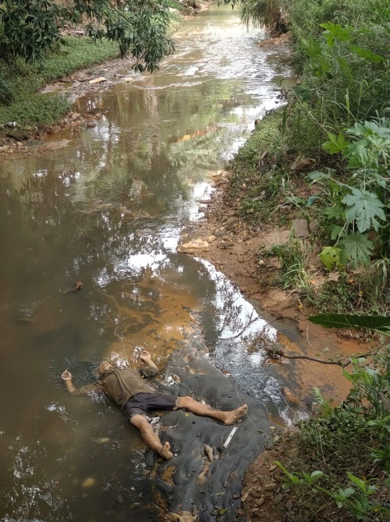 Corpo da vítima as margem do córrego em uma area de mata fechada.