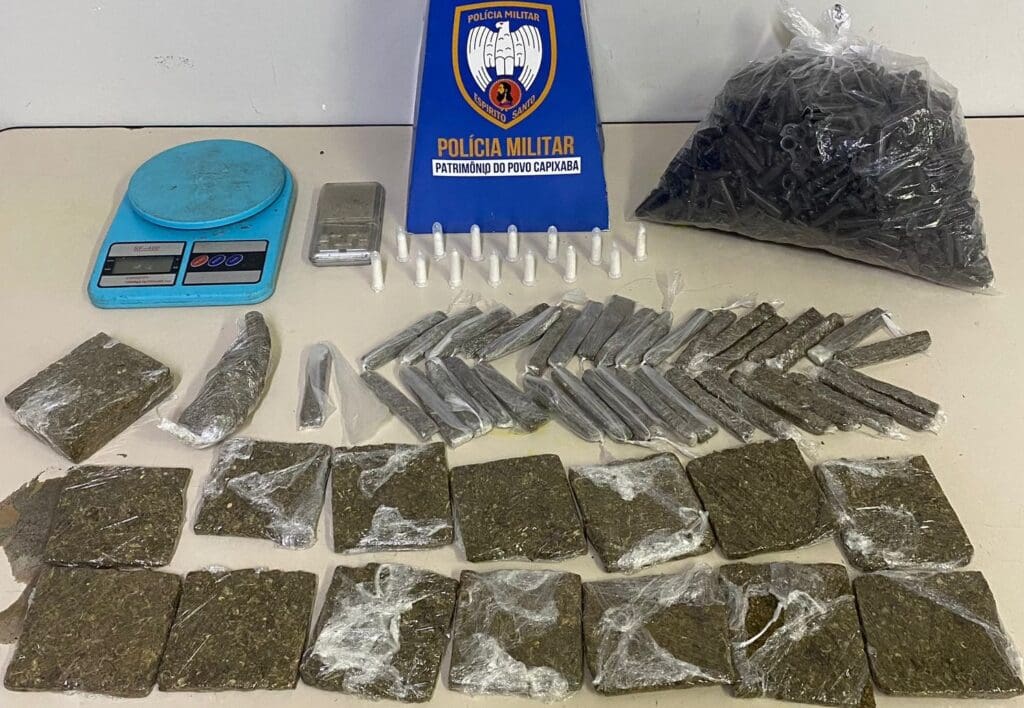 Material apreendido pela Polícia Militar, sobre uma mesa branca 14 tabletes de maconha, 38 buchas da mesma droga, 14 pinos de cocaína, duas balanças de precisão e uma sacola com pinos vazios destinados à embalagem de entorpecentes.