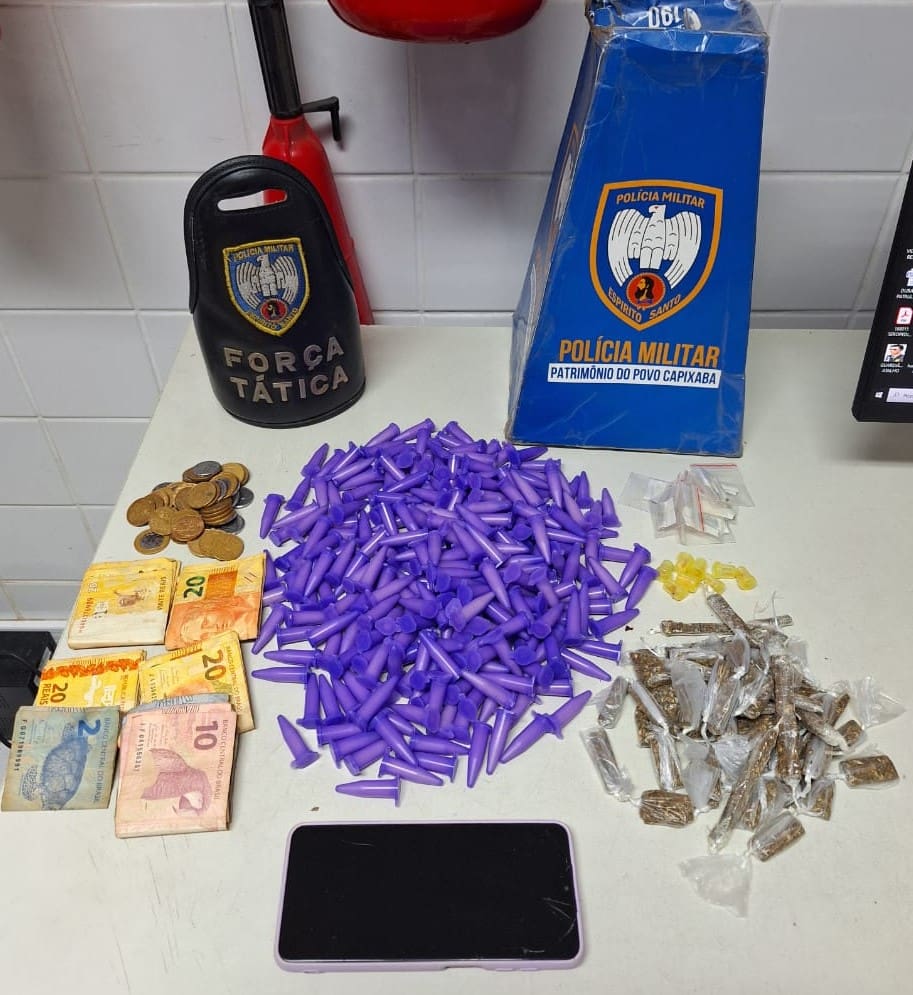 Sobre uma mesa branca, os materiais apreendidos pela equipe da Polícia Militar 292 pinos de cocaína, 43 buchas de maconha, 8 pedras de crack, 8 unidades de haxixe, um celular e R$ 420 em espécie.