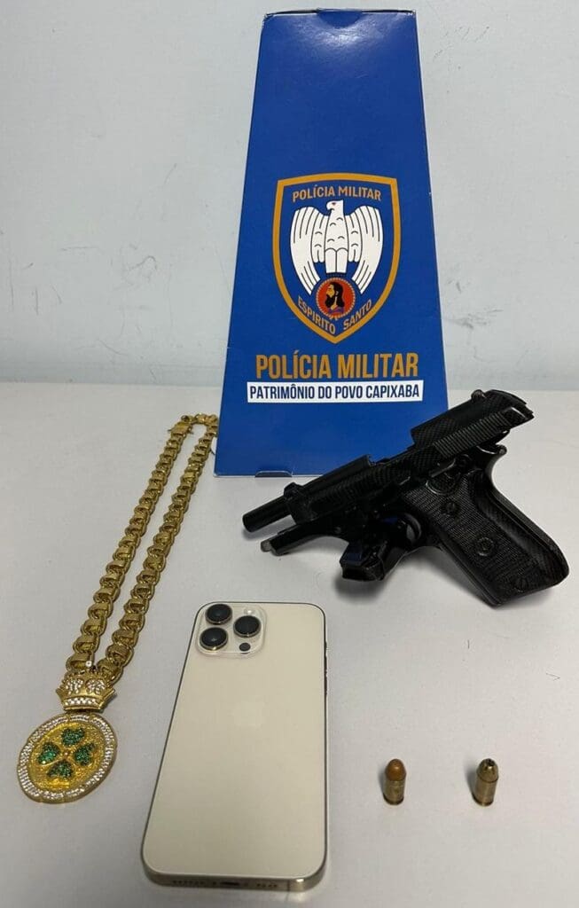 Sobre uma mesa materiais apreendidos pela equipe da Polícia Militar uma pistola calibre .380, duas munições, um celular e uma corrente.