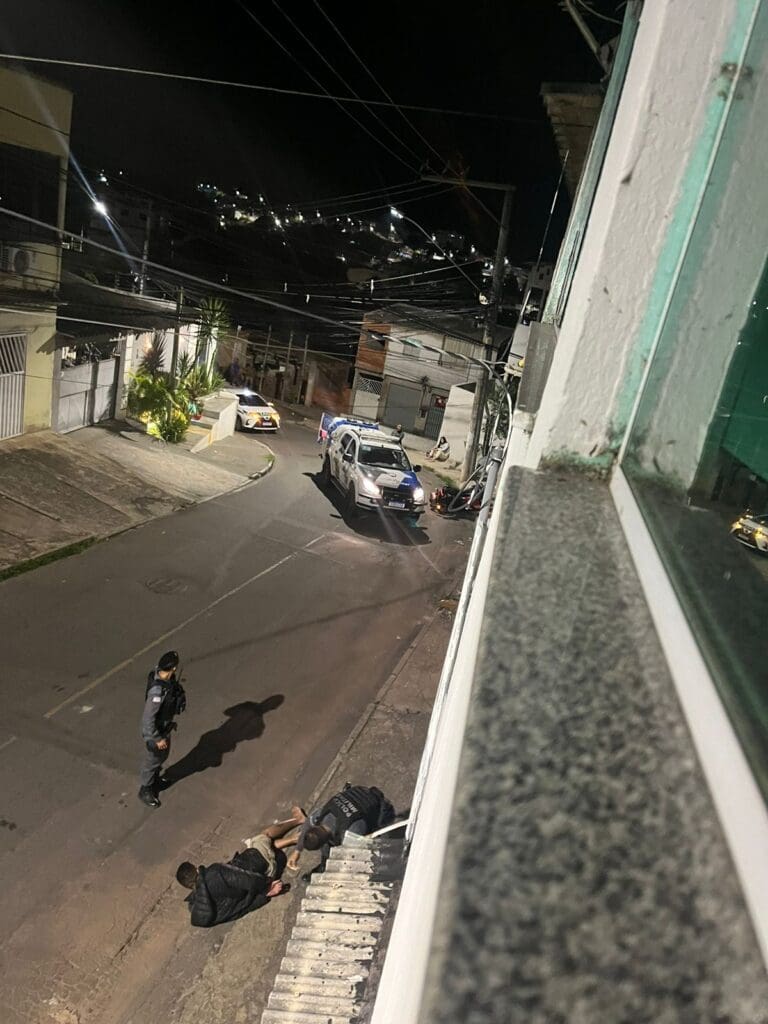 Imagem de uma viatura da Polícia Militar parada no meio de uma rua, e os militares realizando a prisão de um sujeito que esta deitado no asfalto.