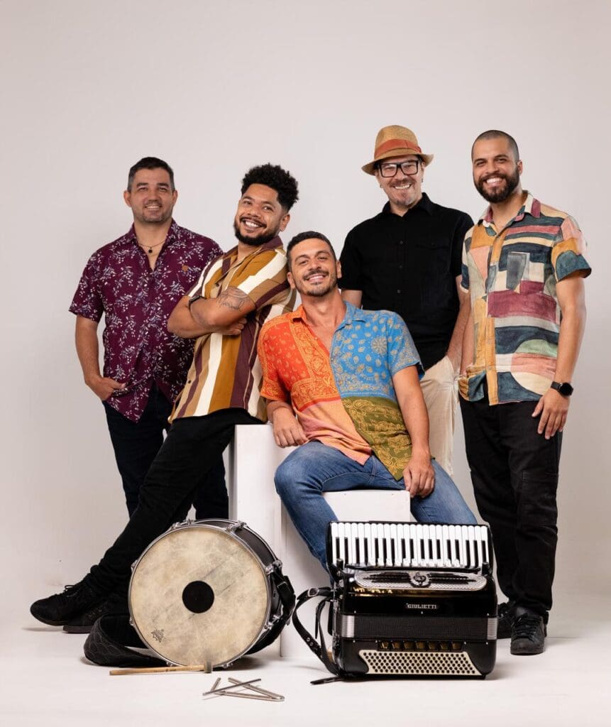 Cinco integrantes da banda: Forró Bem Ti Vi, sorrindo e posando para foto, no chão um tambor, baquetas e uma sanfona.
