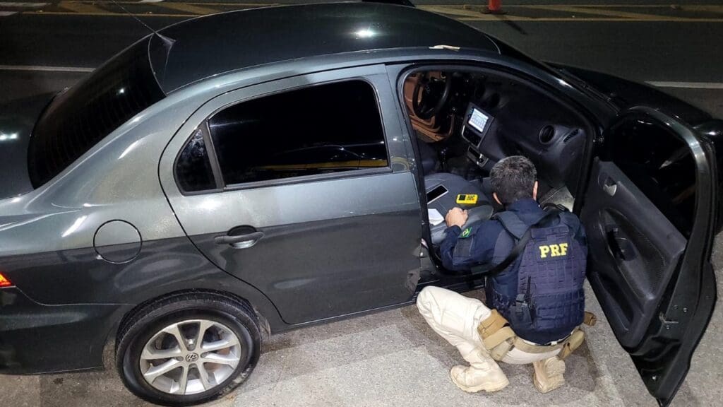 Um agente da Polícia Rodoviária Federal, abaixado ao lado de um carro com uma das portas abertas, vistoriando o veículo recuperado, durante uma abordagem.