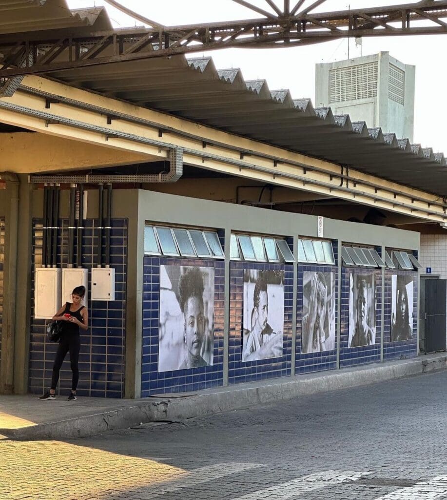 A parede de um terminal de ônibus com 5 painéis com fotos preto e branco de pessoas.