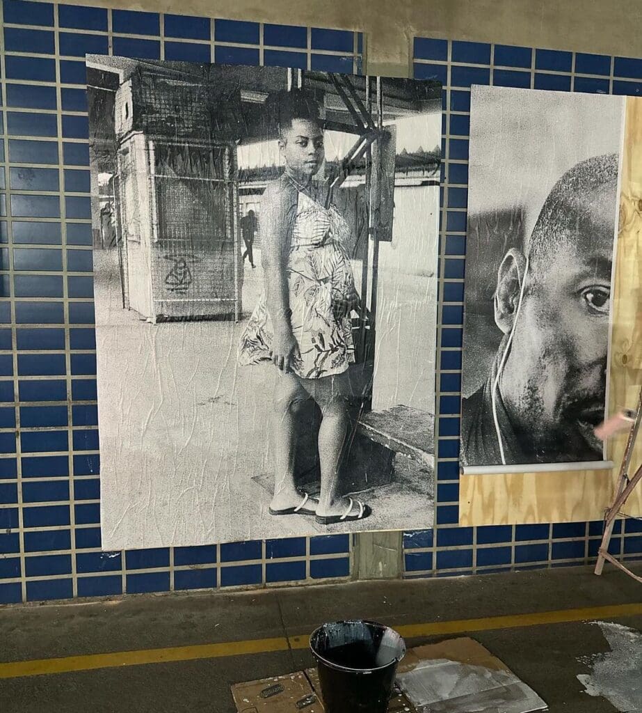 Uma foto preta e branca de uma jovem de vestido gravida ao lado de uma foto de um homem que está sendo colada em um painel, dentro de um terminal de ônibus da Serra.