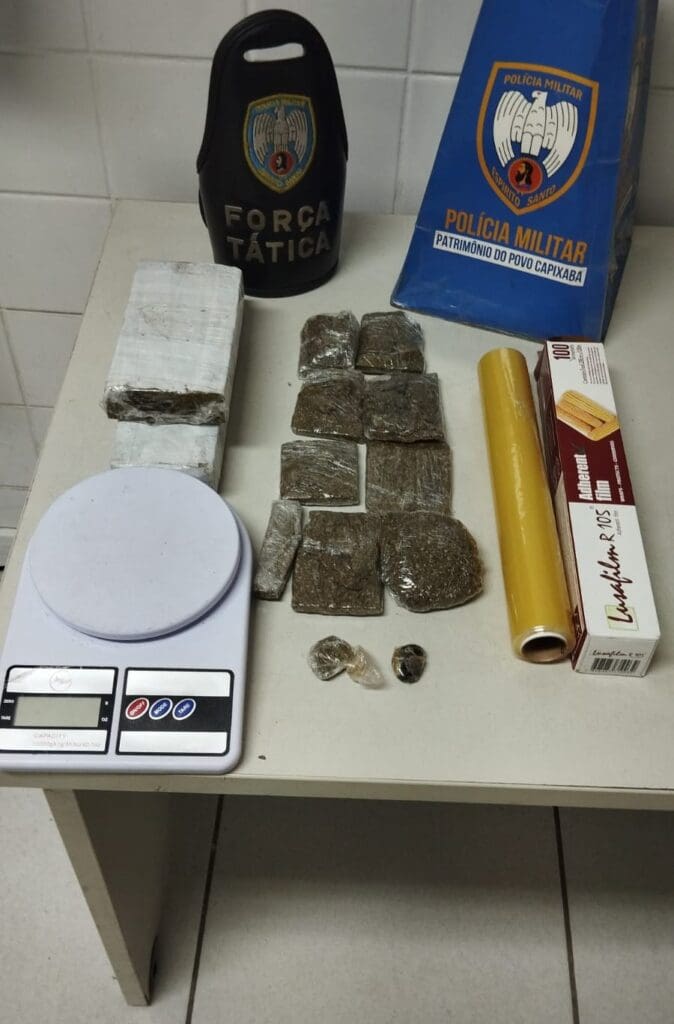 Imagem de uma mesa com uma braçadeira da Força Tática, um cone de papelão usado para divulgar apreensões, dois tabletes de maconha, uma balança de precisão, um rolo de plástico filme além de outros pedaços de maconha.