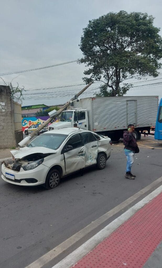 Imagem de um cruzamento, após um acidente, com um carro de passeio amassado, e ao fundo o poste caído sobre o caminhão.