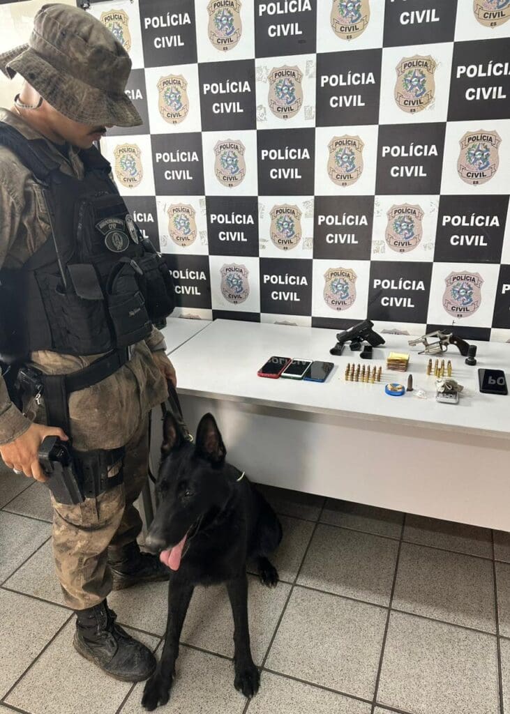 Um policial militar do BAC, junco 
 com um cão farejador ao lado de uma mesa com materiais apreendidos, sobre uma mesa celulares, armas, drogas, munições além de dinheiro apreendidos durante a operação da Polícia Civil.  