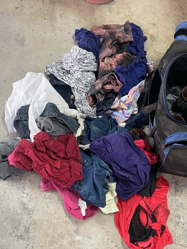 Várias peças de roupas, no chão, ao lado de uma bolsa, os itens são pertences da vítima encontrados na casa do acusado.