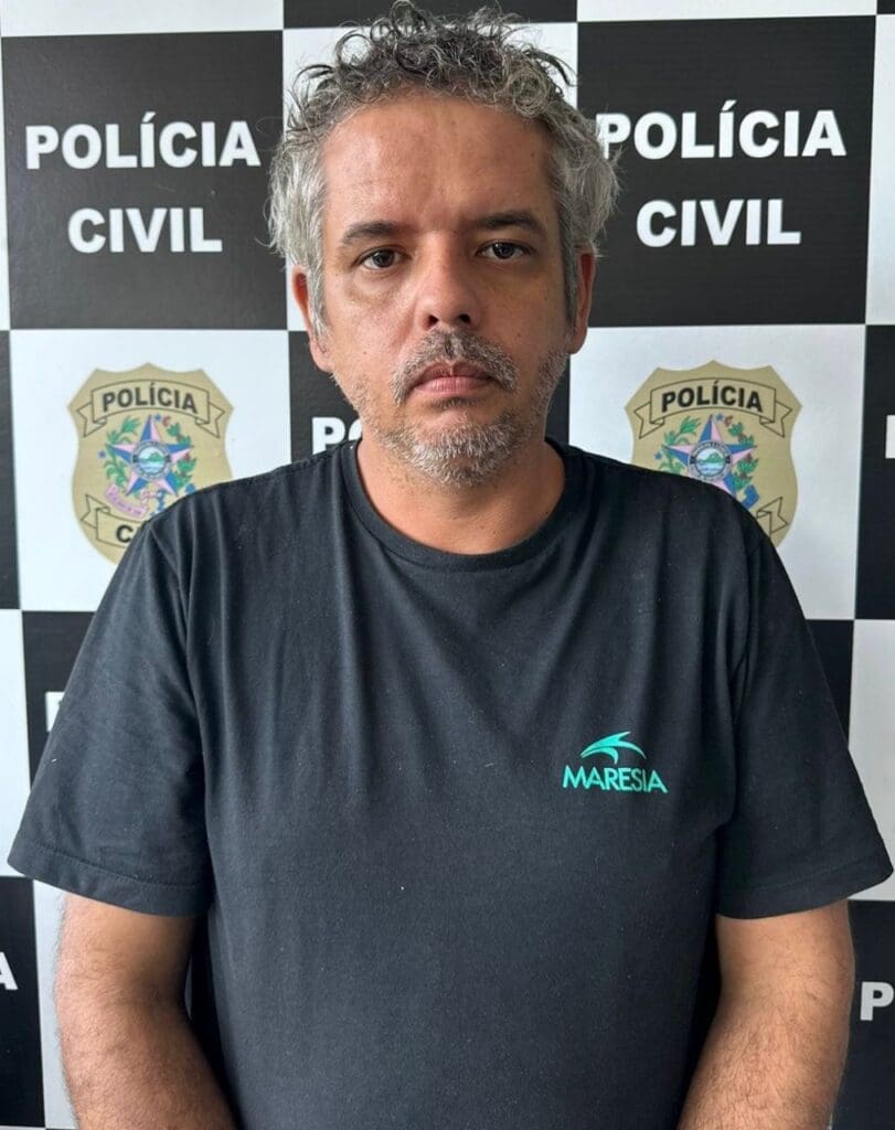 Um homem (Emerson Fontoni de 32 anos), responsável pela morte de um jovem, posando para a foto; no fundo uma parede xadrez com a logo da Polícia Civil, intercalando com o nome.