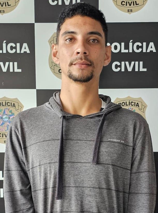 Um sujeito em pé a frente do banner da Polícia Civil após ser preso, pelo assassinato de um motorista.