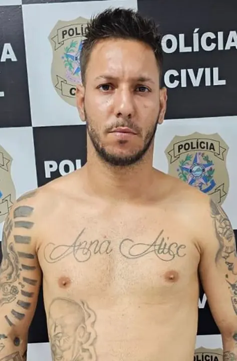 Imagem de um homem identificado como sendo Tiago dos Santos, posando para a foto em frente ao banner da Polícia Civil, após ser preso.