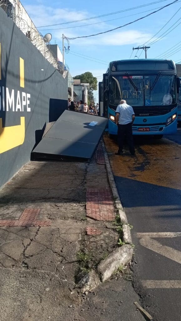 Imagem de um abrigo de passageiros destruído após um acidente com um coletivo, ao lado da estrutura o ônibus e o motorista.