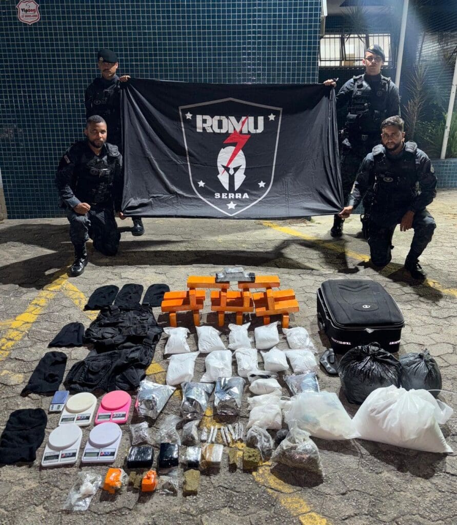 4 agentes da ROMU segurando uma bandeira com o símbolo da corporação, e sobre o chão diversos materiais apreendidos.