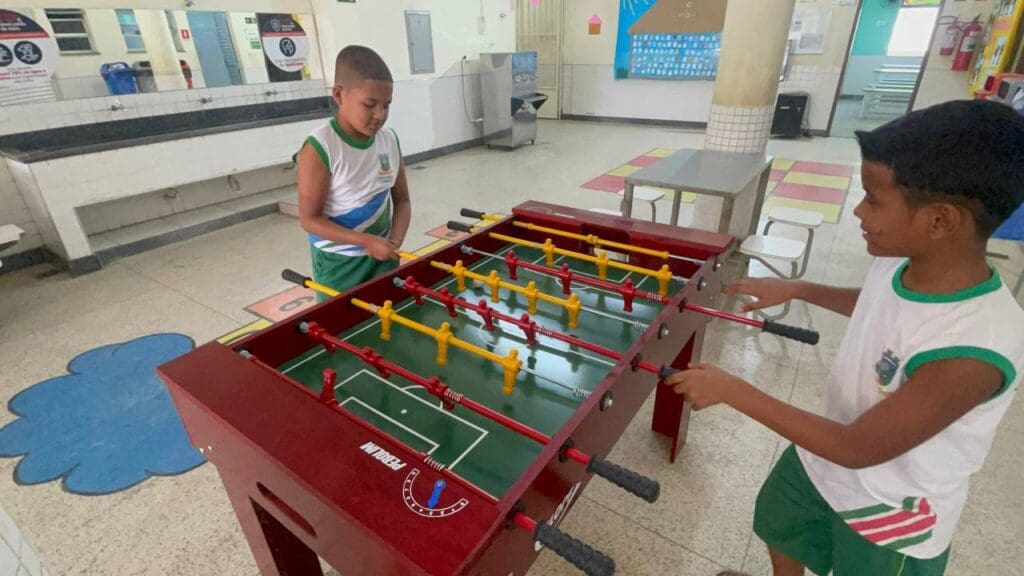 Duas crianças jogando pebolim em um patio de uma escola. 