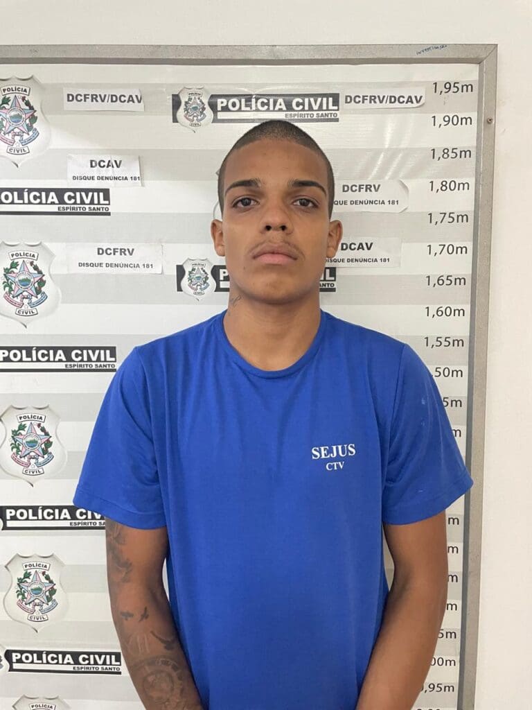 Um homem encostado em uma parede com informações da Polícia Civil e uma marcação de altura, usada para determinar a altura dos presos, (Maycon Vinícius Santos Silva, de 21)
