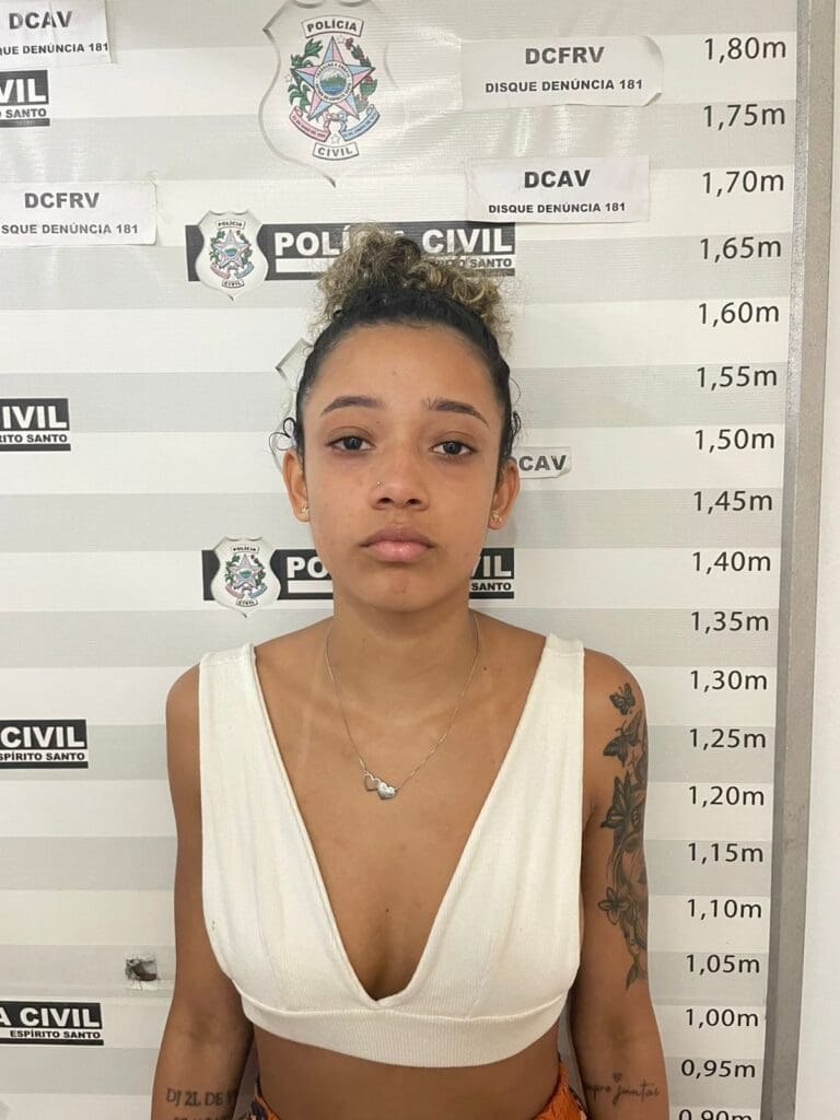 Uma mulher encostada em uma parede com informações da Polícia Civil e uma marcação de altura, usada para determinar a altura dos presos, (Ana Rilery Chaves Alves, de 19 anos)