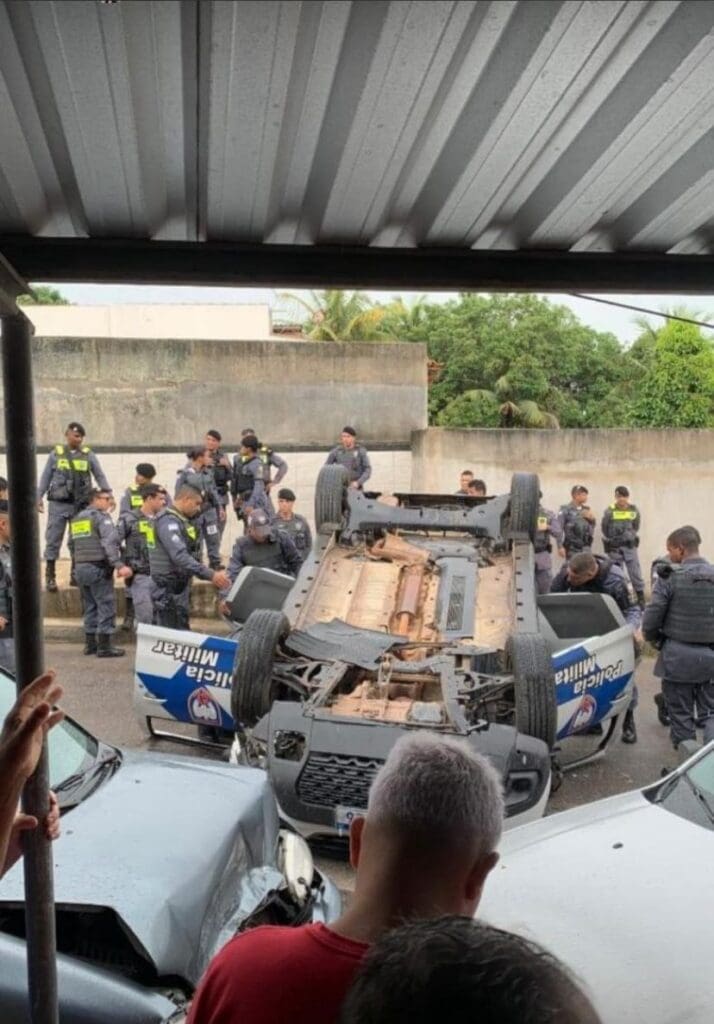 Uma viatura da Polícia Militar capotada em uma rua com diversos policiais militares em volta, proximo a calçada outros dois veículos amassados, que foram atingidos pela viatura. 