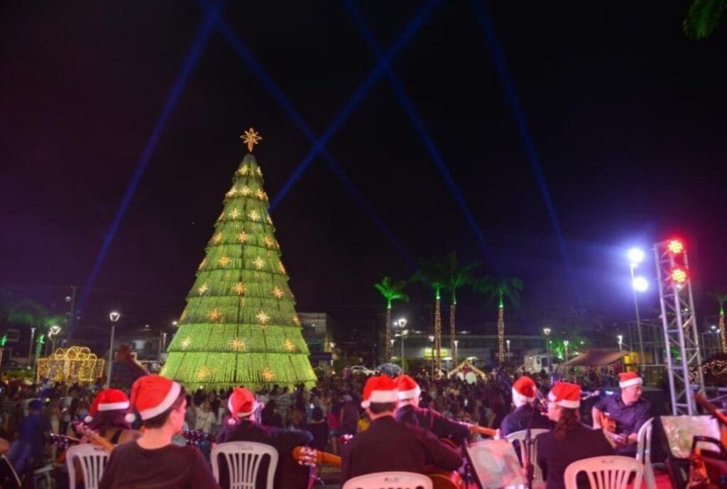 Imagem de um parque com decorações natalinas e uma árvore de Natal ao centro, com as luzes acesas, em volta diversos músicos se apresentando com chapeis de Natal.