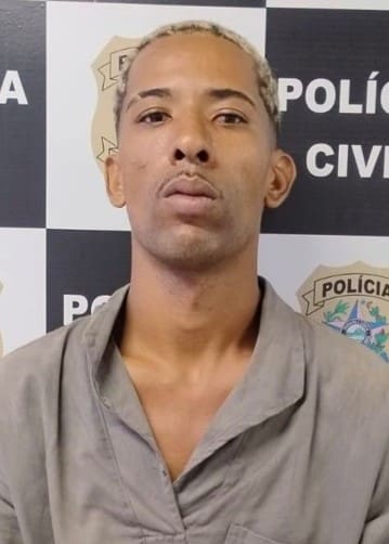 Foto de um jovem, (Junio Silva de Souza, de 26 anos) após ser preso pela Polícia Civil em frente a uma parede com divulgações da Polícia Civil.