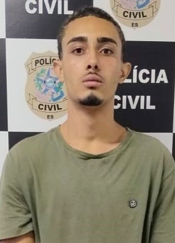 Foto de um jovem, (Endley de Souza Marciel, 19 anos) após ser preso pela Polícia Civil em frente a uma parede com divulgações da Polícia Civil.